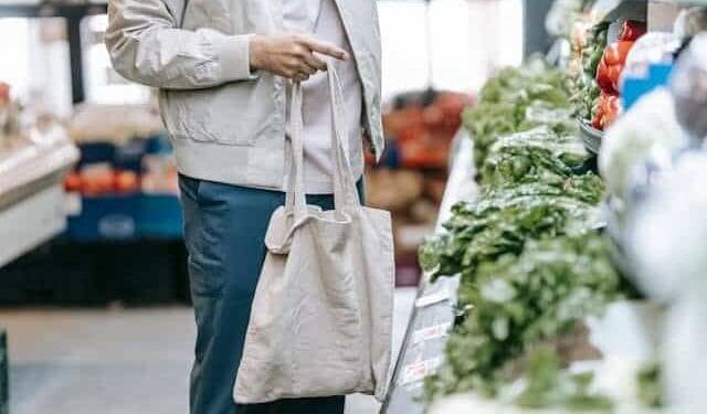 grocery-shopping Mass deportation man holding a reusable shopping tote looking at green vegetables in a grocery store.