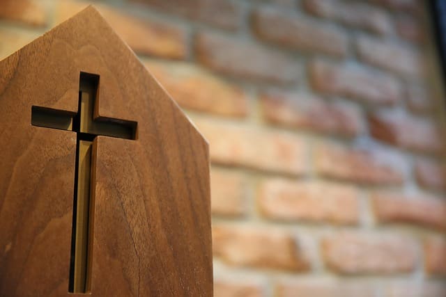christian nationalist cross image by 성원 조 A cross carved into wood with a brick wall behind it.